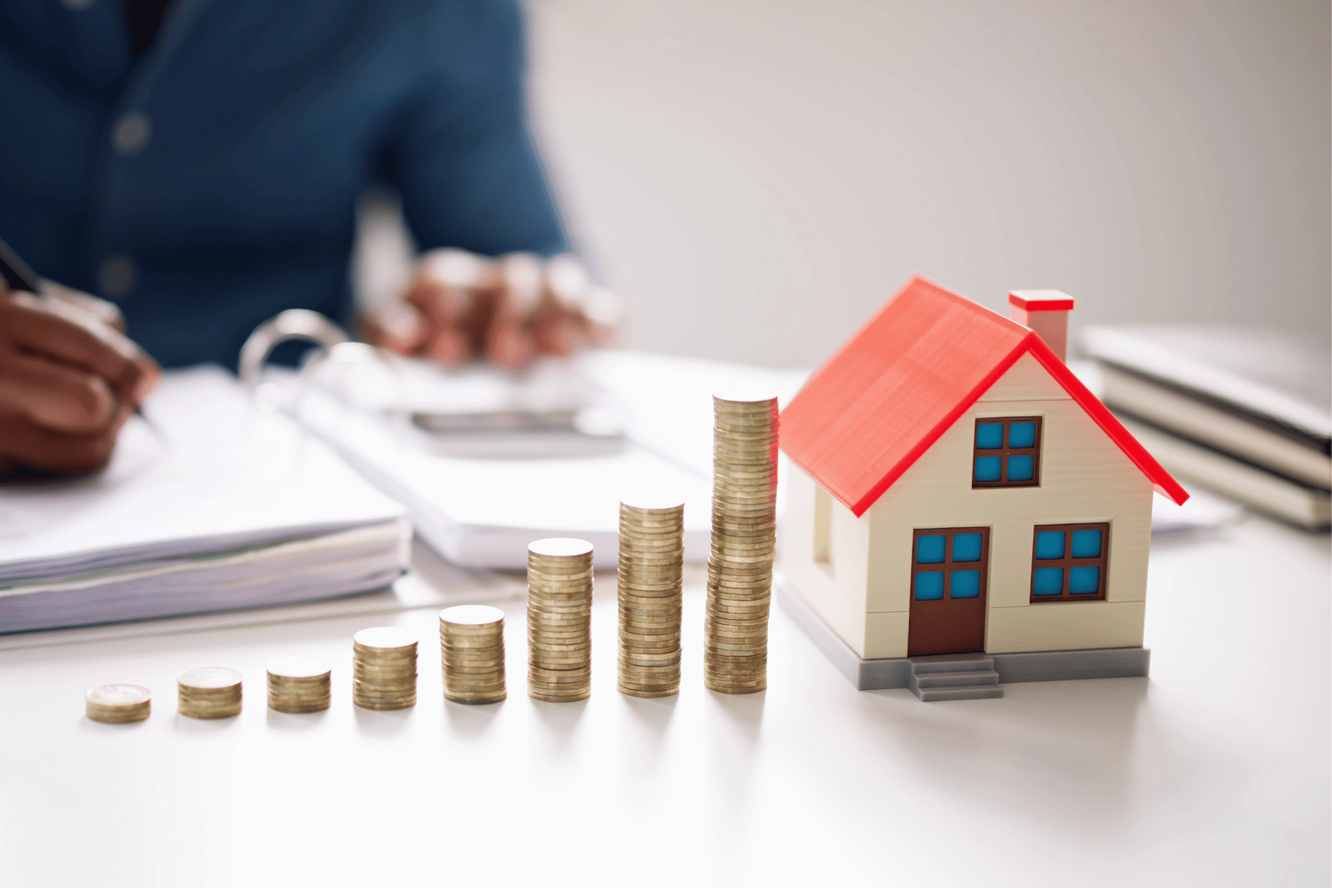 Im Hintergrund sitzt eine Person und schreibt. Im Vordergrund steht rechts ein kleines Modellhaus und von links nach rechts sind höher werdende Münzentürme gestapelt.