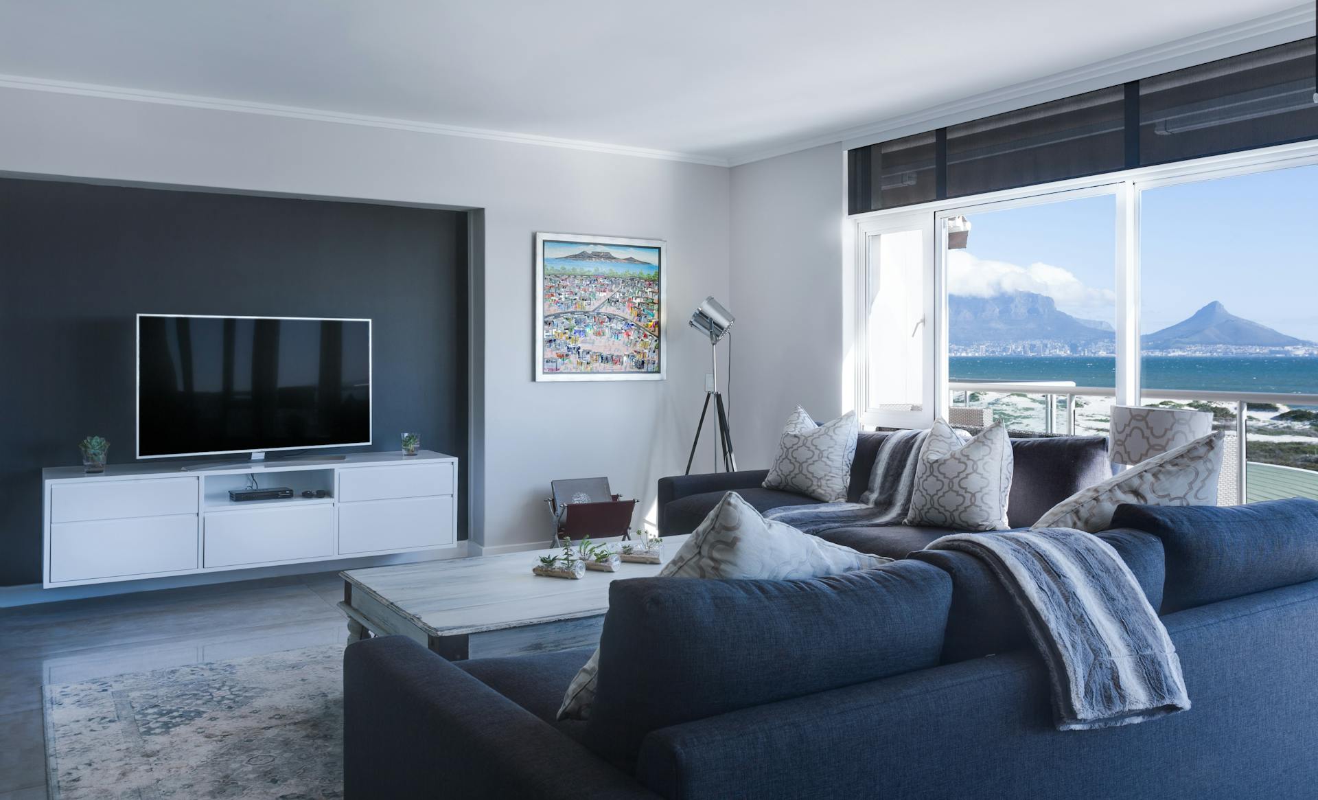 Bild eines modern eingerichteten Wohnzimmers. An der Wand hängt ein Fernseher. Die Wände sind weiß und dunkelgrau gestrichen. Rechts ist ein Fenster. Im Vordergrund ist eine dunkelblaue Couch zu sehen. 