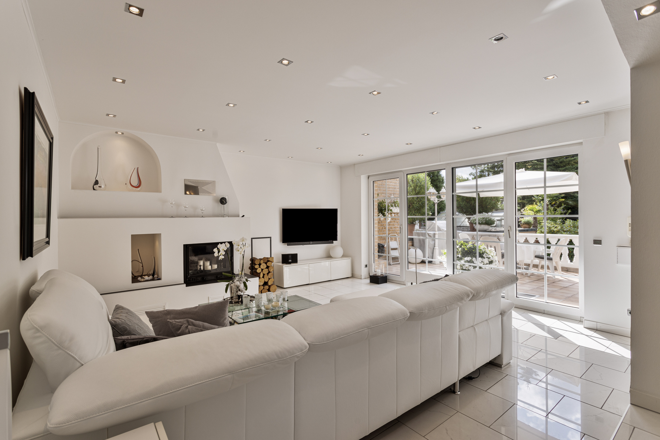 Weißes Wohnzimmer mit einer großen weißen Couch. Rechts im Bild sieht man die Terrasse mit Gartenmöbeln. 